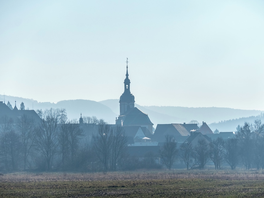 A church with a tall steeple, surrounded by trees and buildings in a foggy landscape.
