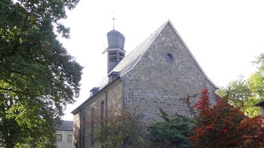 A stone church with a bell tower, surrounded by trees and red flowers.