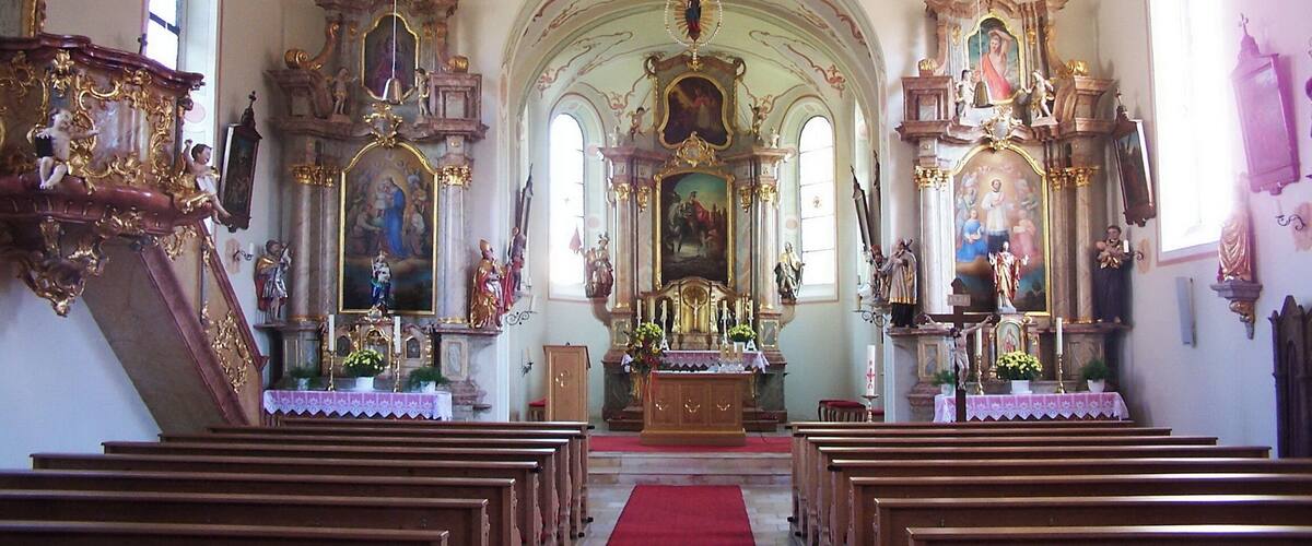 An ornate church interior with wooden pews, a red carpet, and elaborate religious paintings and statues.