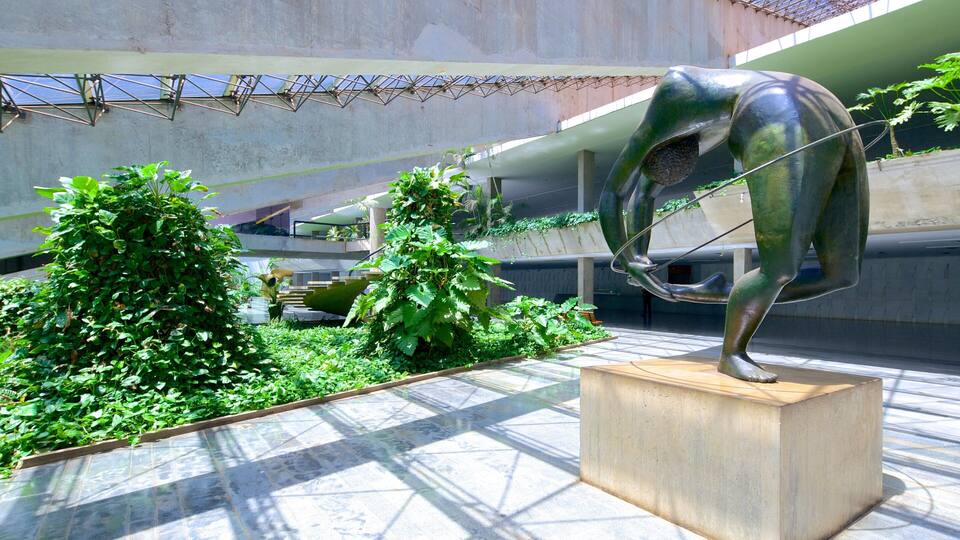 Escultura de una figura en una pose dinámica, rodeada de exuberante vegetación y arquitectura moderna.