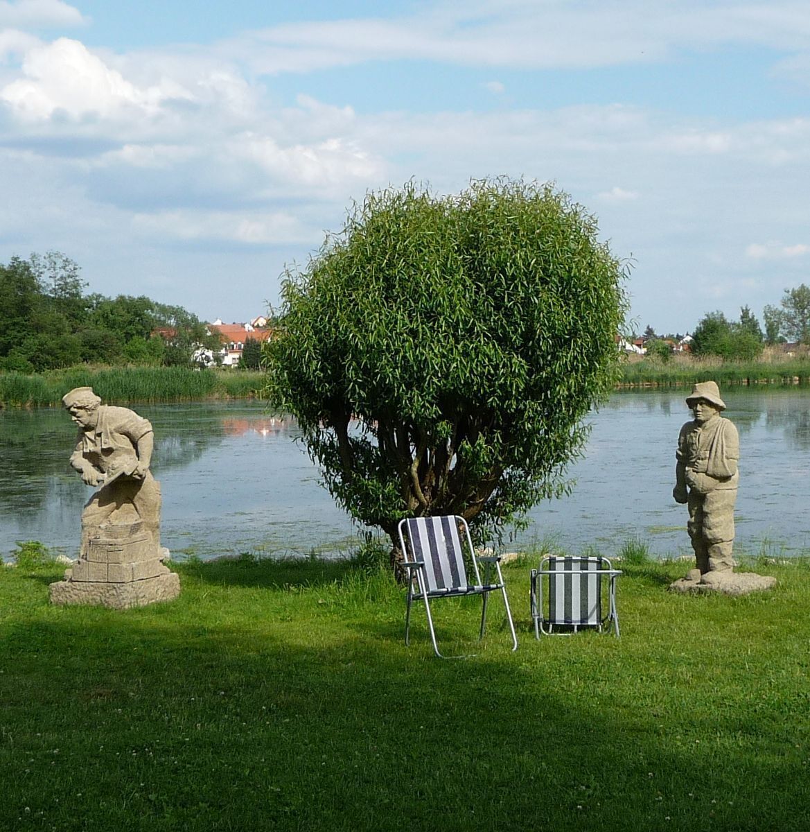 A serene lakeside scene with a large weeping willow tree, two stone statues, and a striped chair by the water.