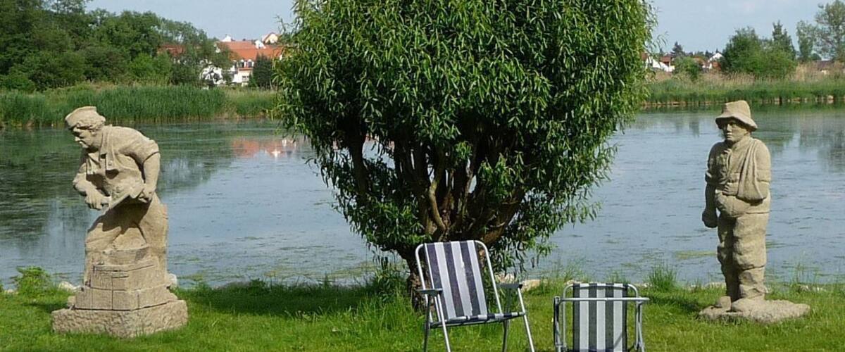 A serene lakeside scene with a large weeping willow tree, two stone statues, and a striped chair by the water.