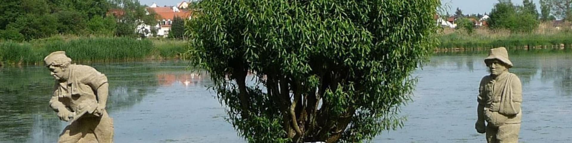 A serene lakeside scene with a large weeping willow tree, two stone statues, and a striped chair by the water.