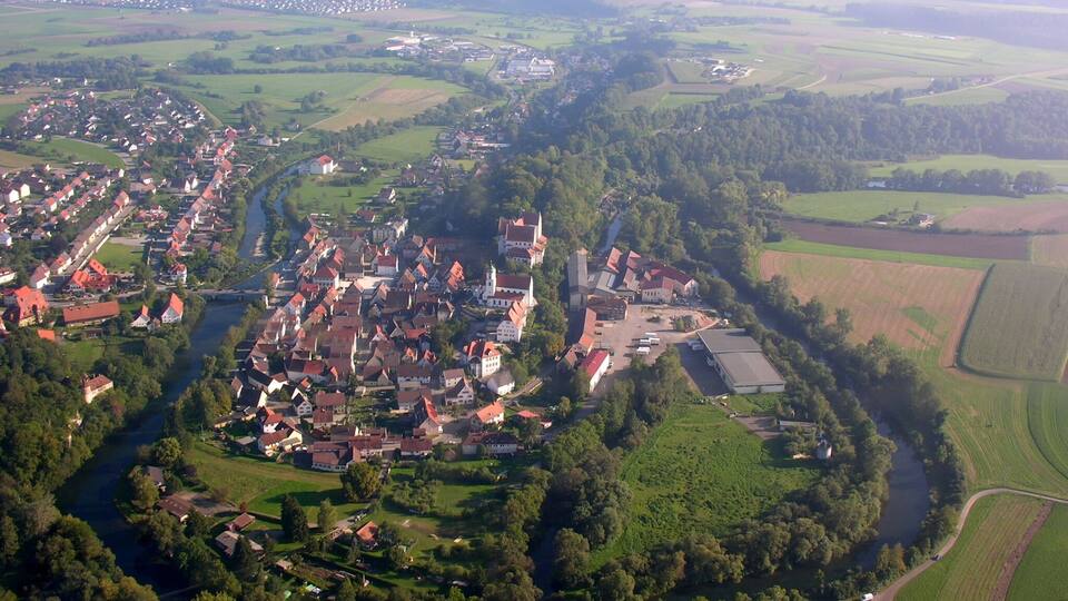An aerial view of a town with a river, surrounded by green fields and a large church.