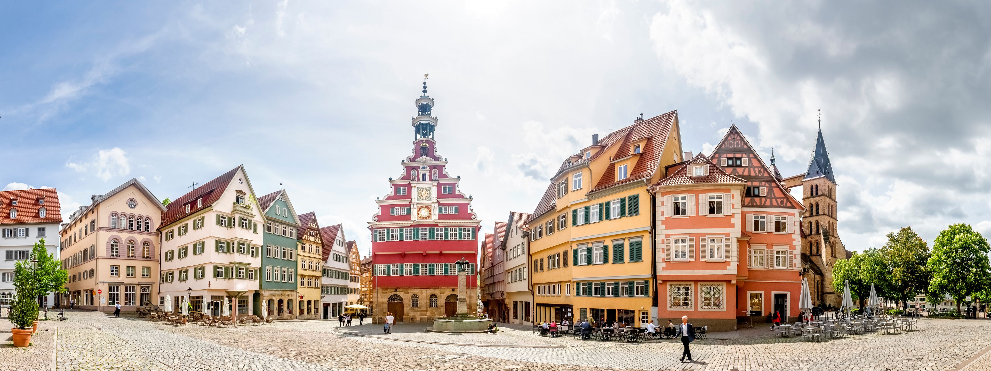 Une place pavée, bordée de bâtiments historiques, avec une tour de l’horloge au centre et des espaces aménagés pour s’asseoir à l’extérieur.