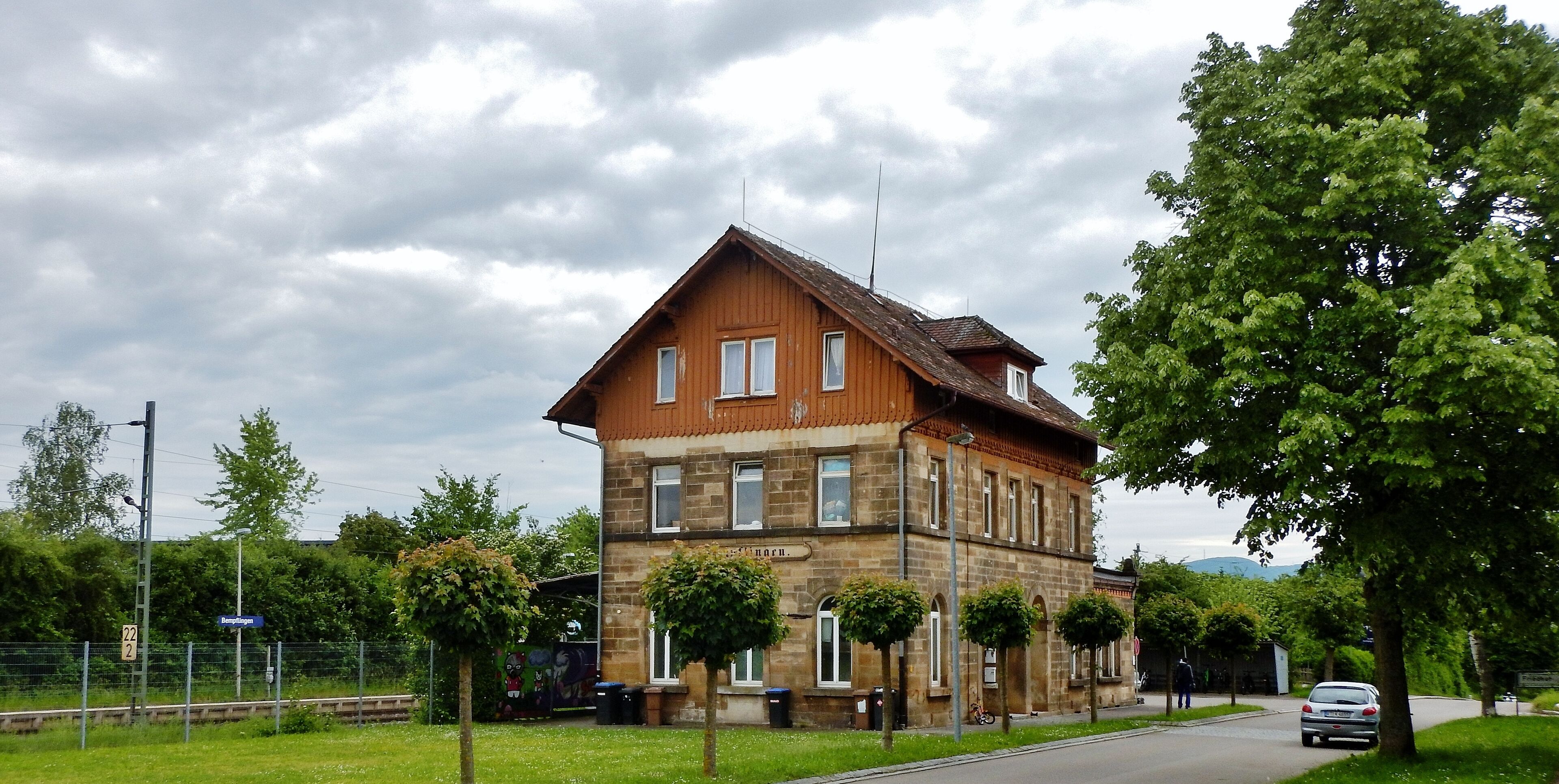 A two-story stone building with a sloped roof, surrounded by trees and a road.