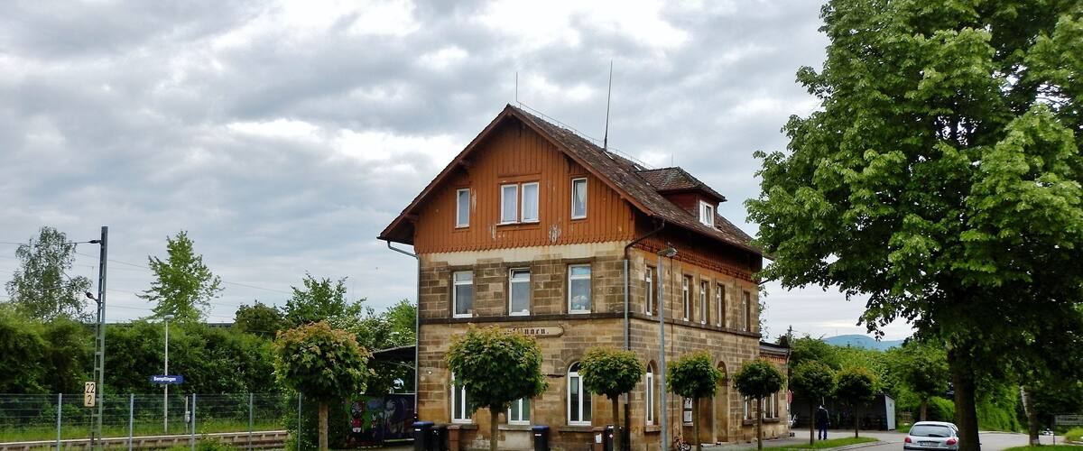 A two-story stone building with a sloped roof, surrounded by trees and a road.