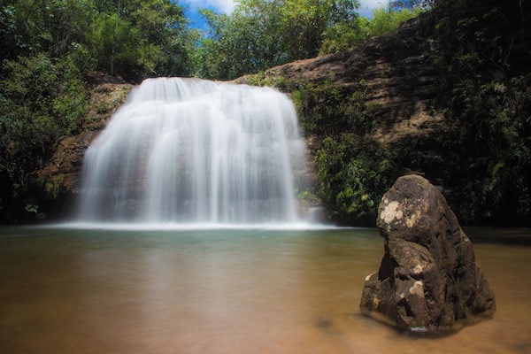 Uma cascata desce sobre um penhasco rochoso, rodeada por vegetação exuberante.