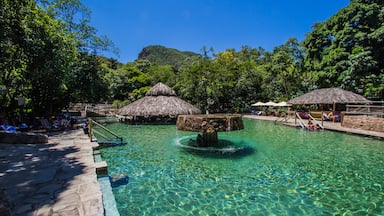 Piscina translúcida com borda de pedra, rodeada por vegetação exuberante e estruturas com telhados de palha.