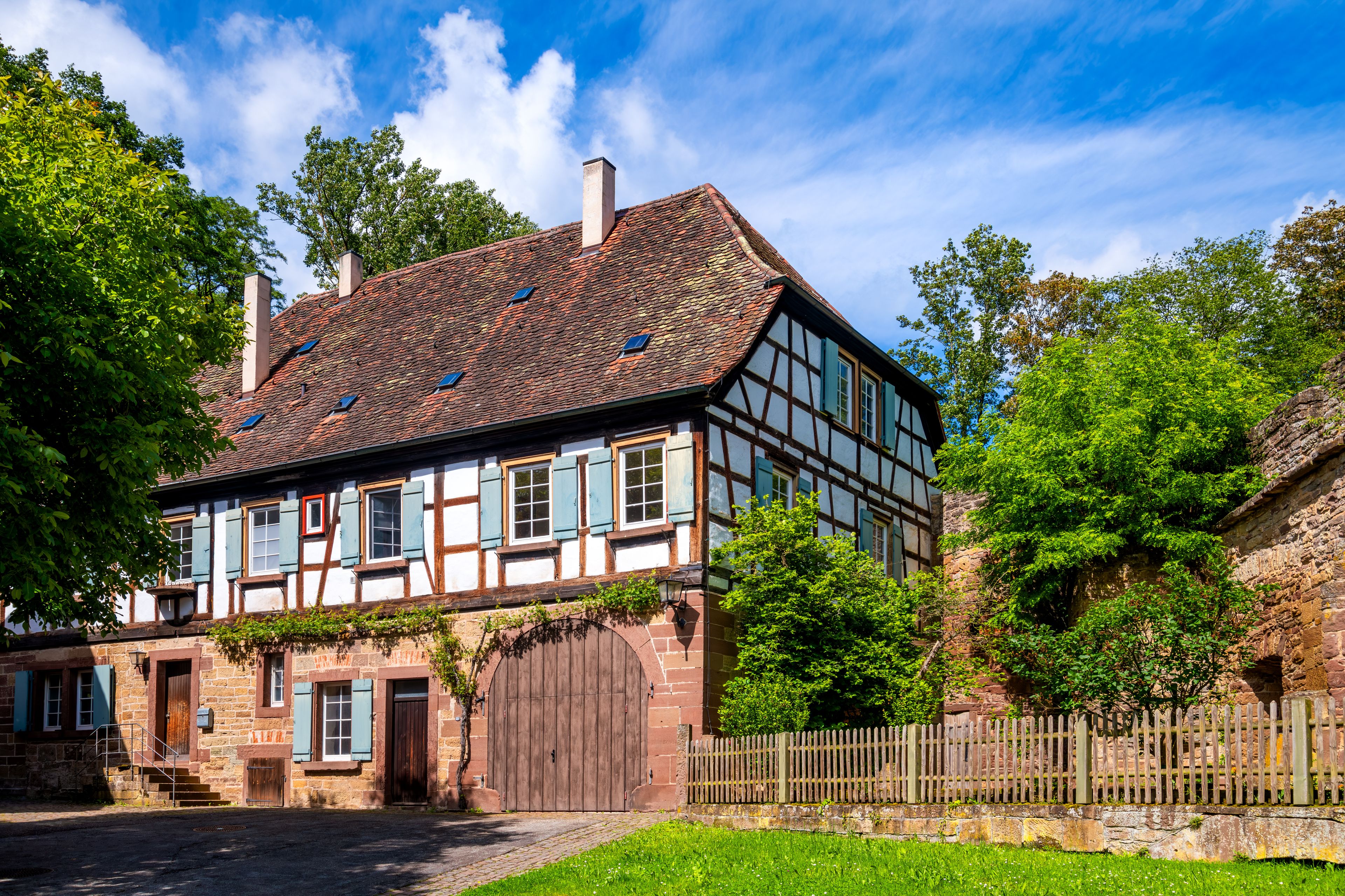 A traditional half-timbered house with a sloped roof, surrounded by lush greenery and a stone wall.