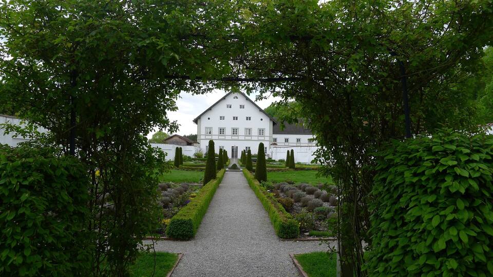 A white building with a symmetrical facade, surrounded by a gravel pathway, and framed by lush green trees.