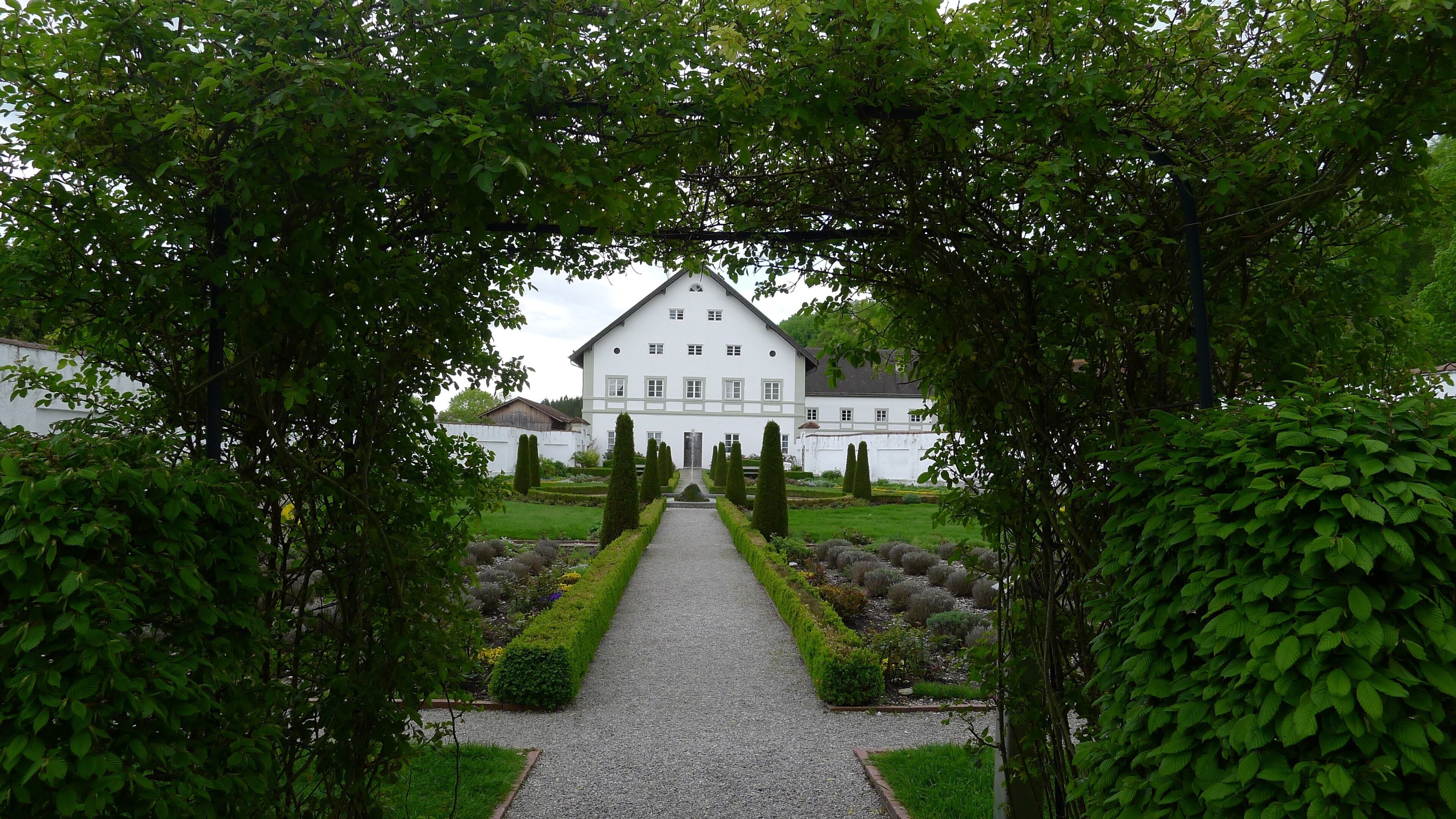 A white building with a symmetrical facade, surrounded by a gravel pathway, and framed by lush green trees.