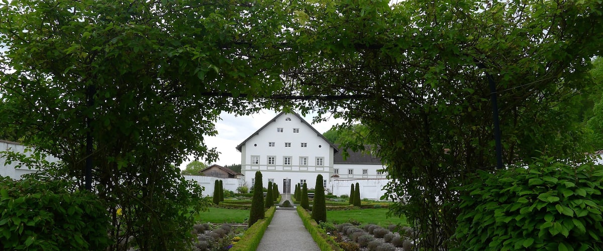 A white building with a symmetrical facade, surrounded by a gravel pathway, and framed by lush green trees.