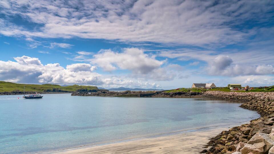 A coastal scene with a boat on calm waters, a sandy beach, rocky shoreline, and a small settlement with buildings.