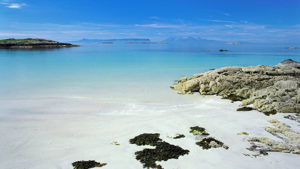 A beach with clear turquoise water, white sand, and rocky outcrops.
