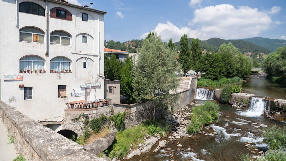 Ein Fluss fließt durch eine Stadt, gesäumt von einer Steinbrücke und einem Gebäude mit Bogenfenstern.