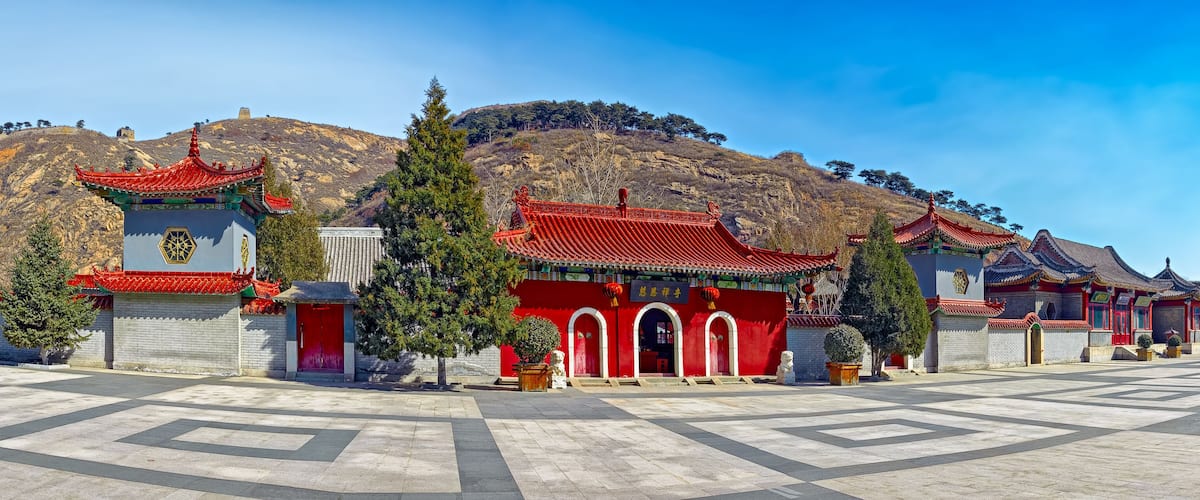 A traditional Chinese architectural complex with red-roofed buildings, a large open courtyard, and a mountainous backdrop.