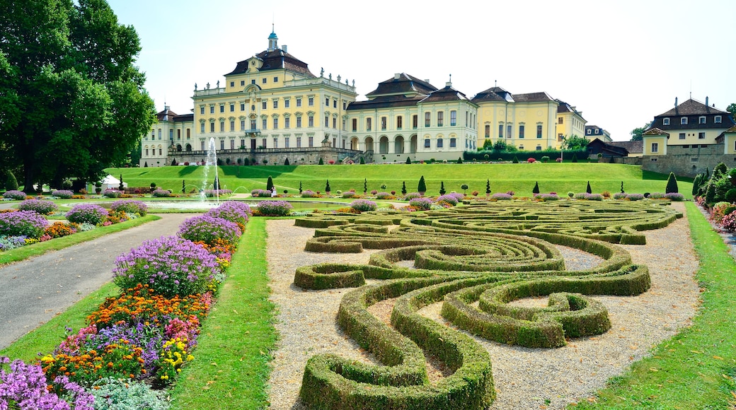 A formal garden with a hedge maze, colorful flower beds, and a large classical building with a clock tower.