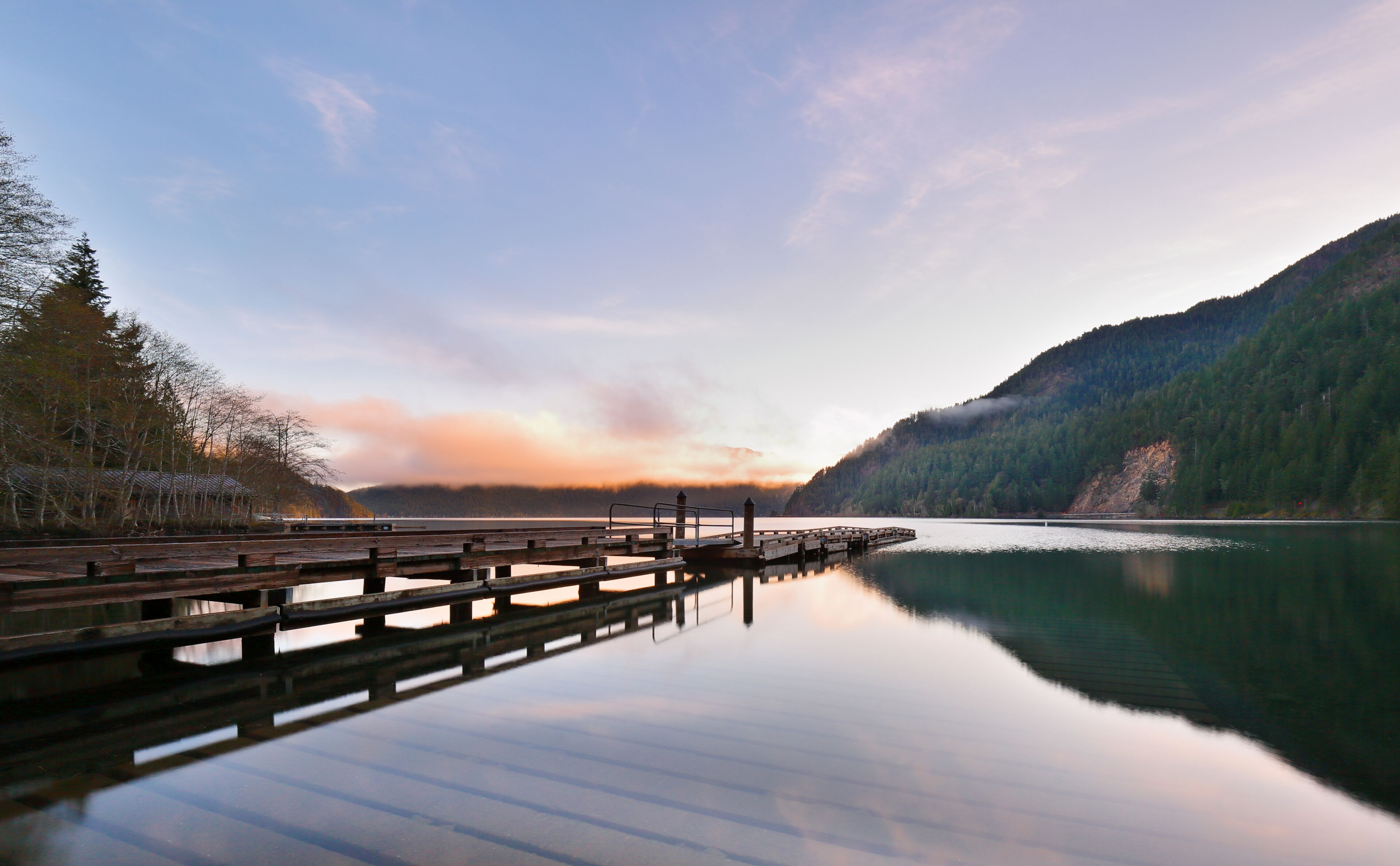 A calm lake with a wooden dock extending into the water, surrounded by forested hills and a clear sky.