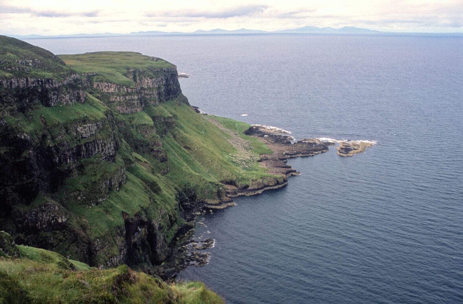 A coastal cliff with green vegetation and rocky outcrops extending into the sea.
