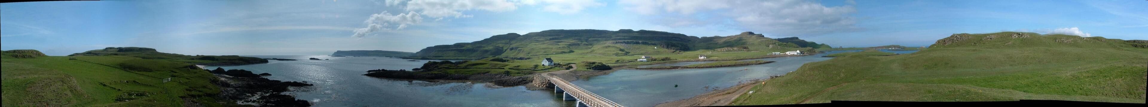 Panorama of the Isle of Canna, outermost of the Small Isles on the west coast of Scotland. Taken from Sanday, looking towards the western, inhabited end of Canna, it shows the bridge linking Canna and Sanday, habitation and the main farm square, Corogan tower in the distance and Canna Bay and the pier to the right hand side.