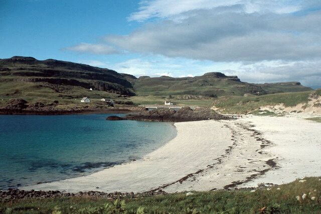 A coastal landscape with a sandy beach, calm waters, rolling hills, and a few scattered houses.