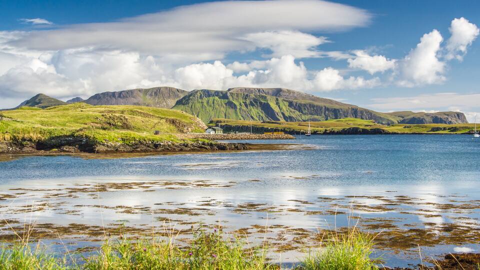 A coastal landscape with green hills, a calm sea, and a small house by the shore.