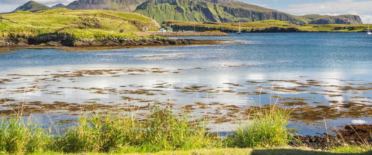 A coastal landscape with green hills, a calm sea, and a small house by the shore.