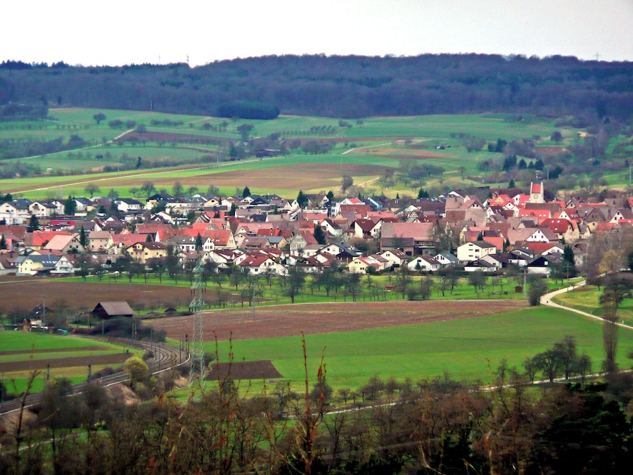 Un pueblo rural con casas de tejados rojos, rodeado de campos verdes y una colina boscosa al fondo.