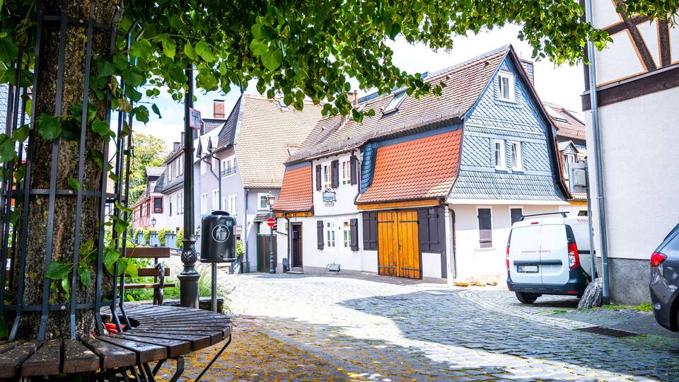 A cobblestone street lined with traditional European buildings, a bench, and a parked van.