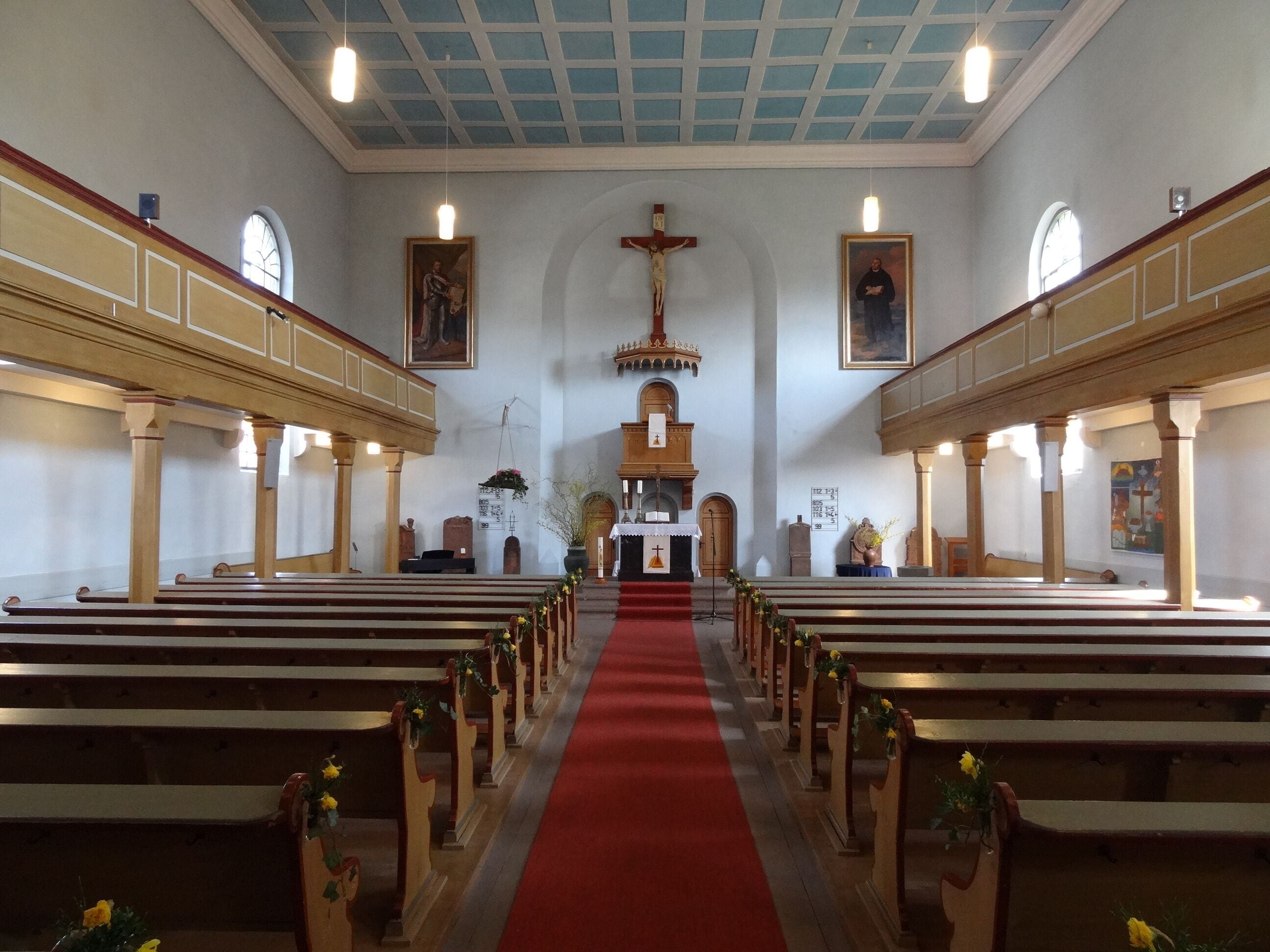 A church interior with wooden pews, a red carpet aisle, and religious paintings on the walls.