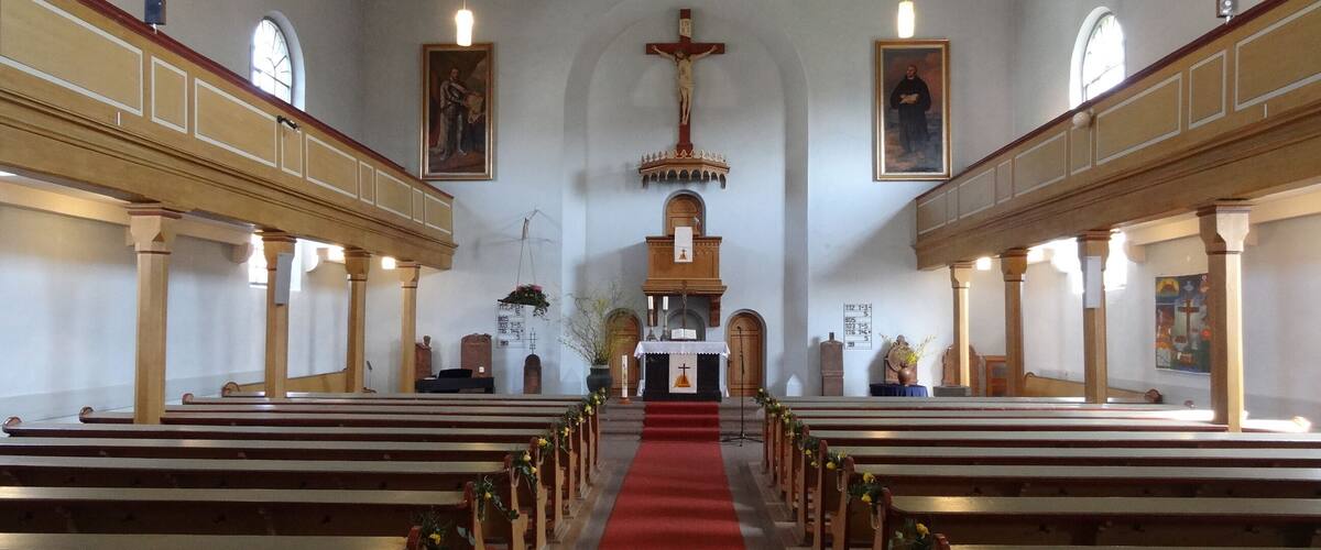 A church interior with wooden pews, a red carpet aisle, and religious paintings on the walls.
