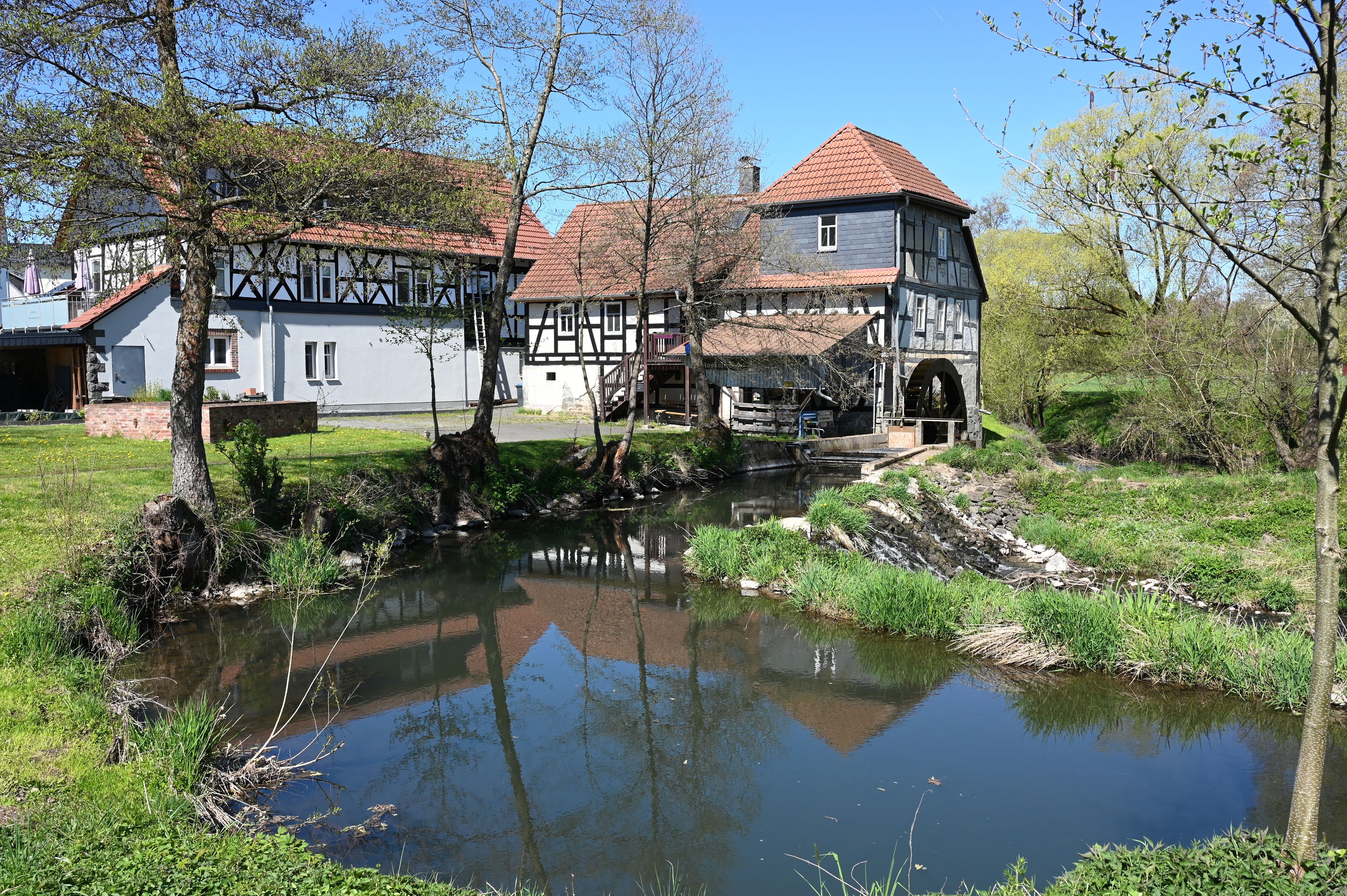 A traditional half-timbered house with a watermill, surrounded by a pond and greenery.