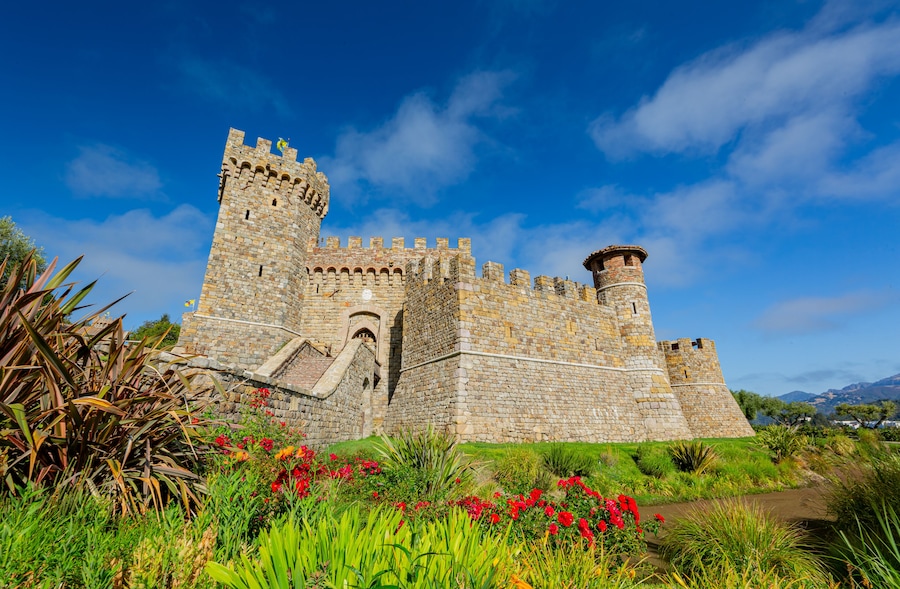 A historic stone castle with a prominent tower, surrounded by lush greenery and vibrant flowers.