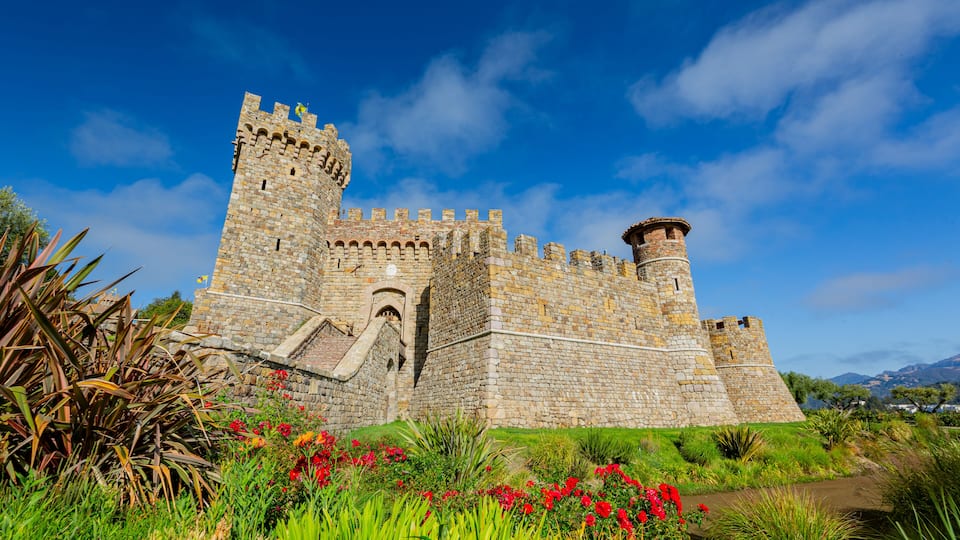 A historic stone castle with a prominent tower, surrounded by lush greenery and vibrant flowers.