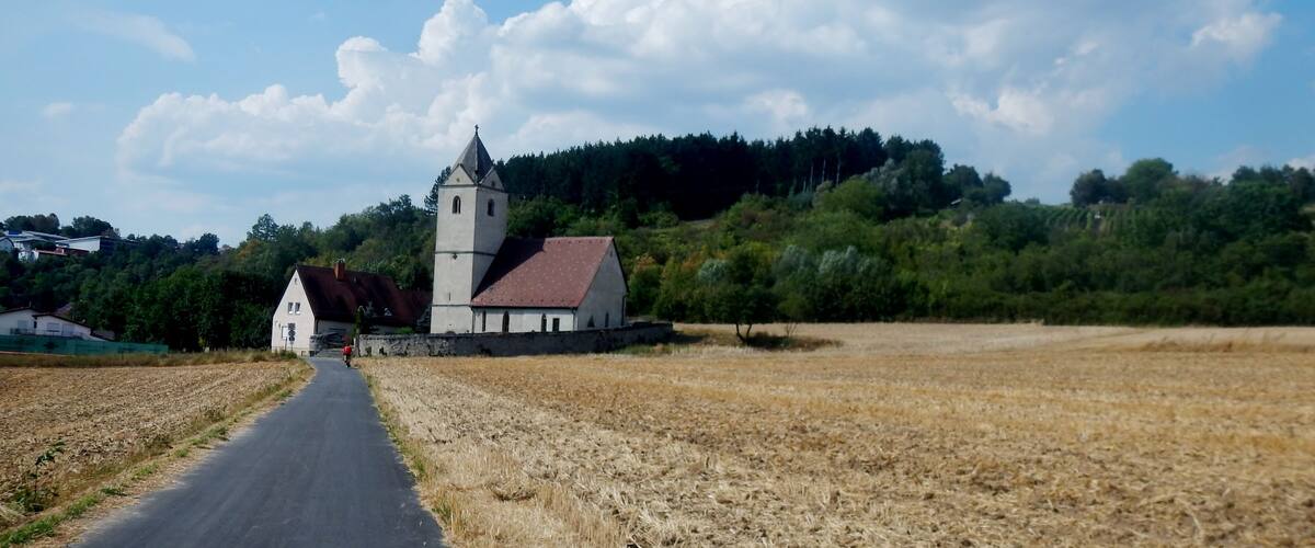 A rural road leading to a church with a tower, surrounded by fields and a forested hill.
