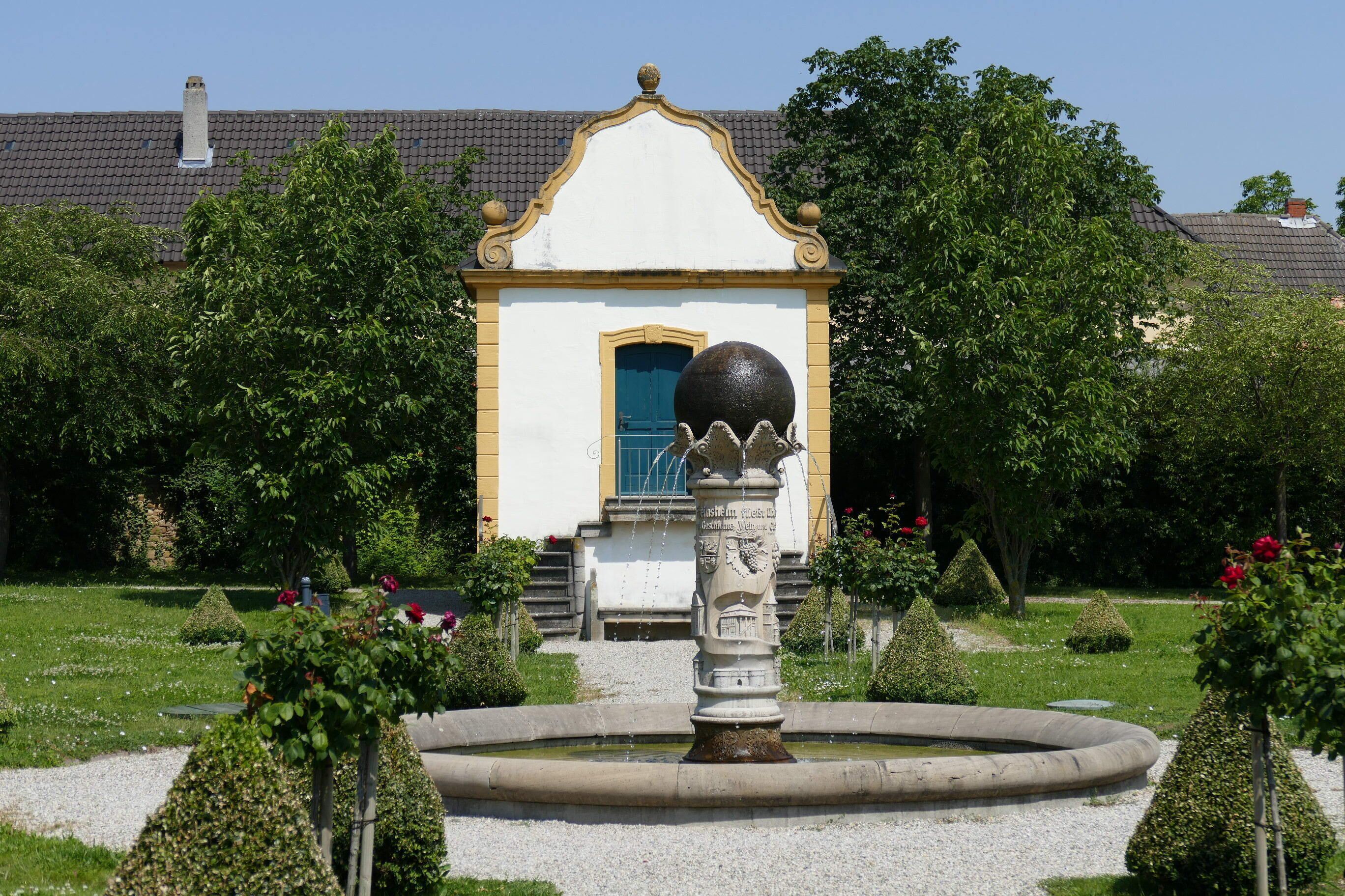 A garden with a fountain, trimmed hedges, and a building with a blue door.