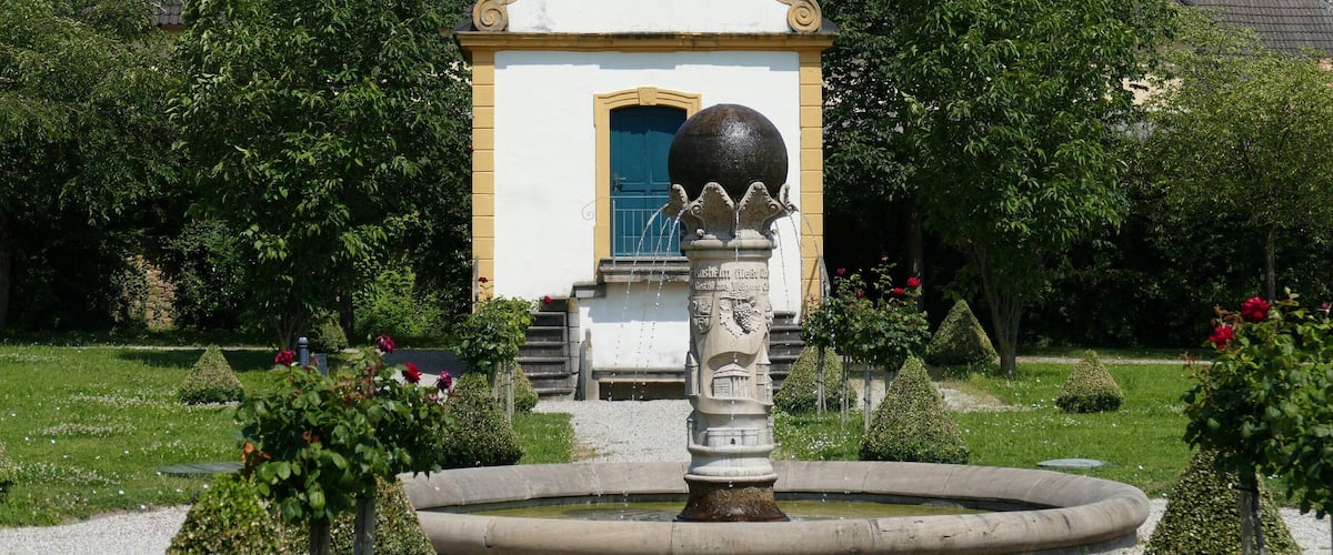 A garden with a fountain, trimmed hedges, and a building with a blue door.