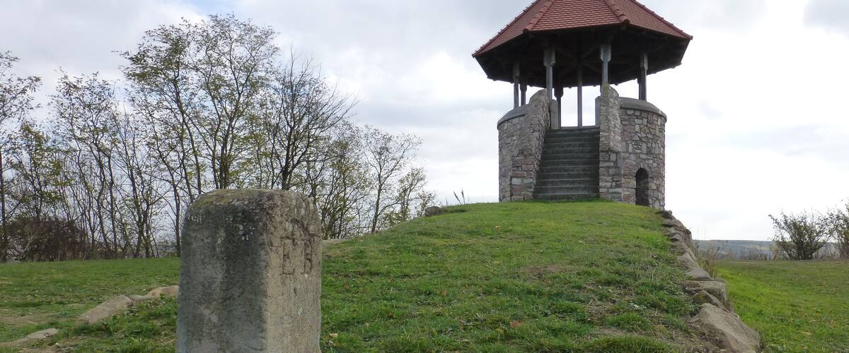 A stone gazebo on a grassy hill with a stone pillar nearby.