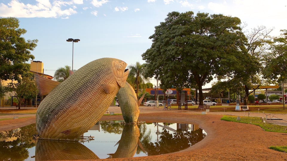 A large fish sculpture in a pond with trees and buildings in the background.