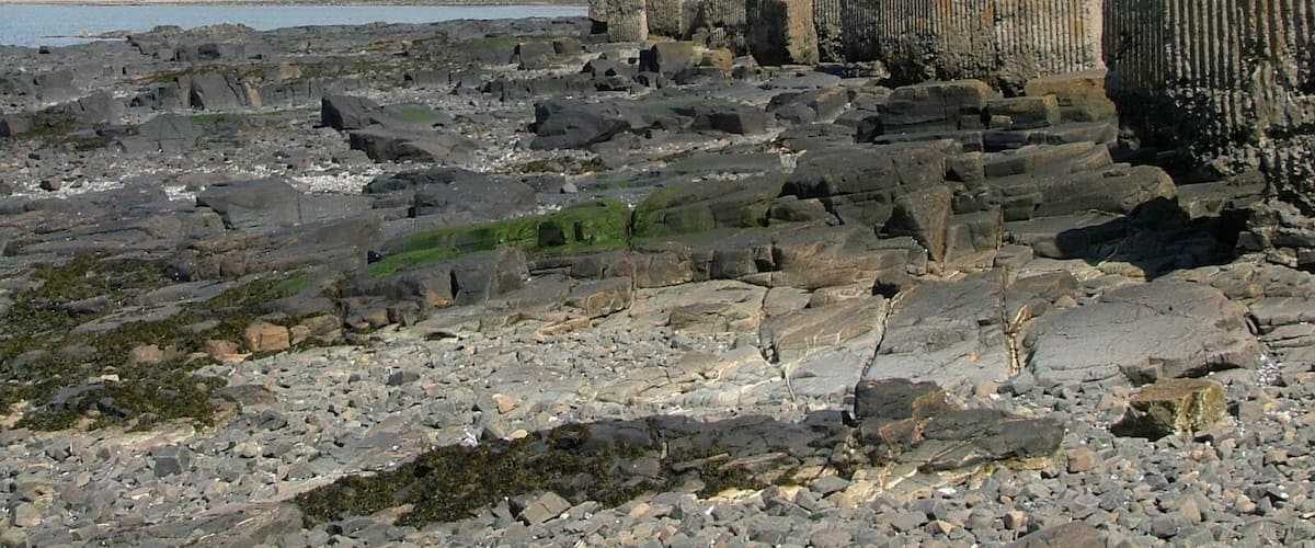 A rocky shoreline with ancient stone structures and a distant building.