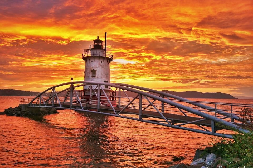 A lighthouse with a metal bridge over water during sunset.