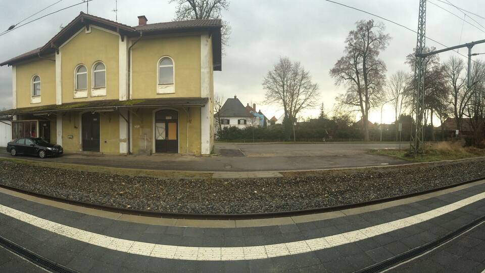A train station platform with a yellow building, a parked car, and a railway track.
