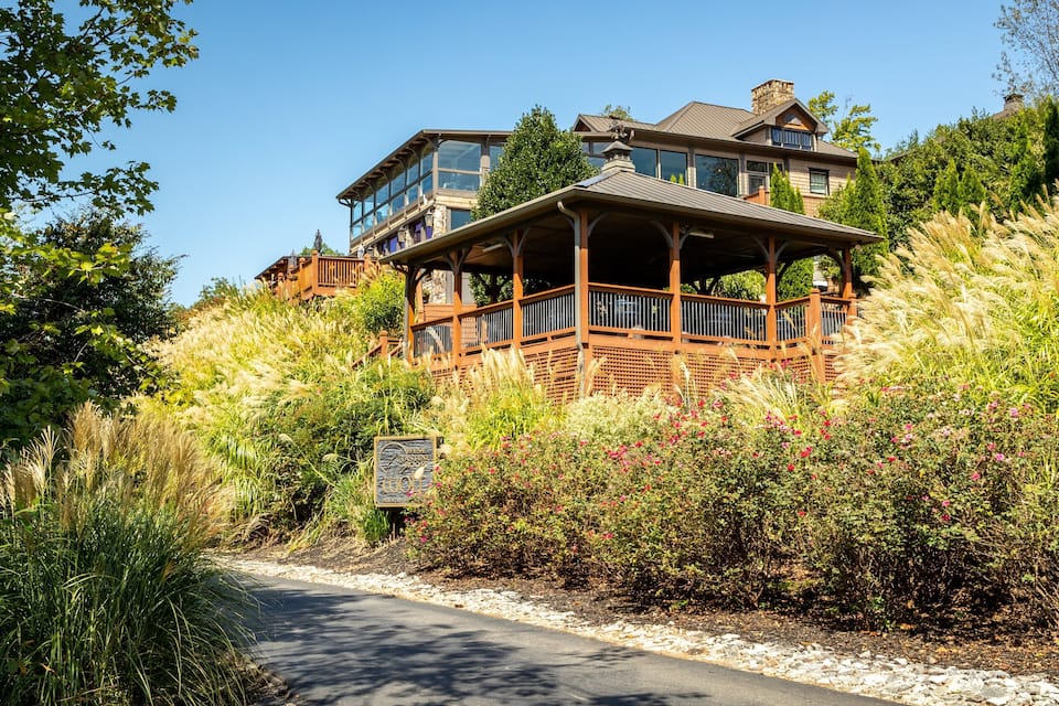 Une grande maison à plusieurs étages, dotée d’un porche en bois et entourée de hautes herbes et de plantes en fleurs.