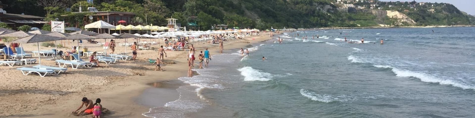 Una playa con gente tomando el sol, nadando y jugando en el agua. Hay sombrillas y sillas de playa en la arena.