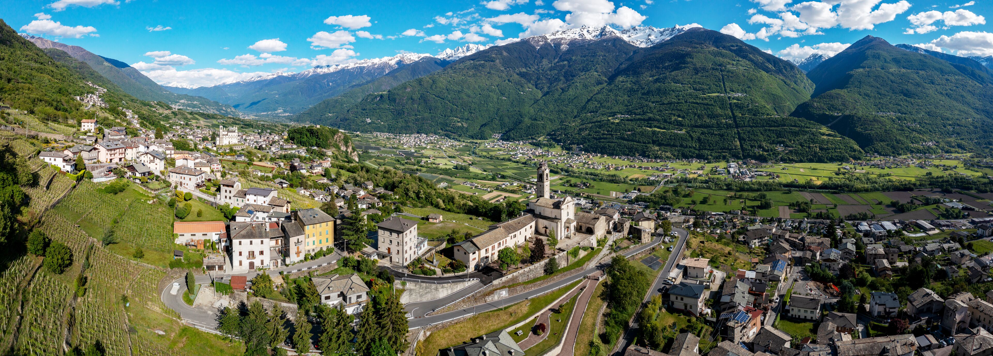A mountainous landscape with a town nestled in a valley, surrounded by lush greenery and snow-capped peaks.