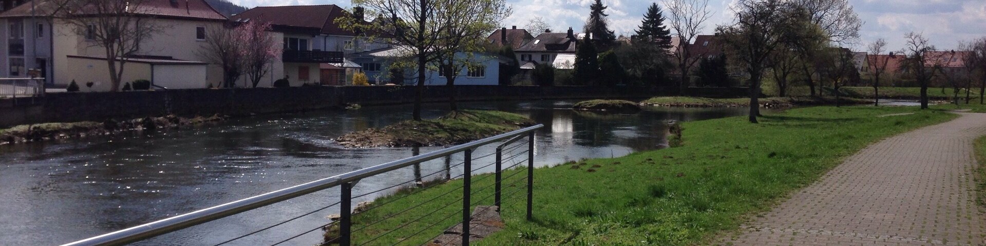 A riverside pathway with a metal railing, a brick walkway, and houses in the background.