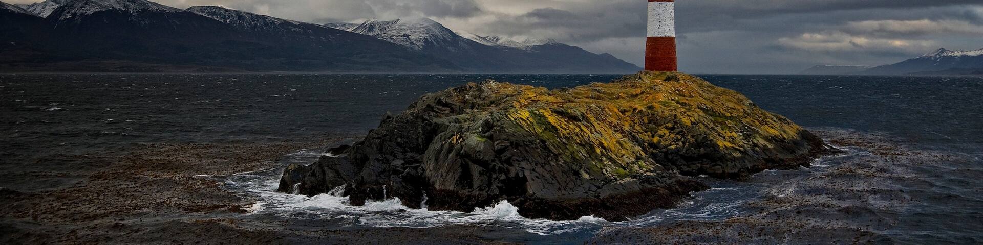 A lighthouse on a rocky outcrop with a backdrop of mountains and a cloudy sky.