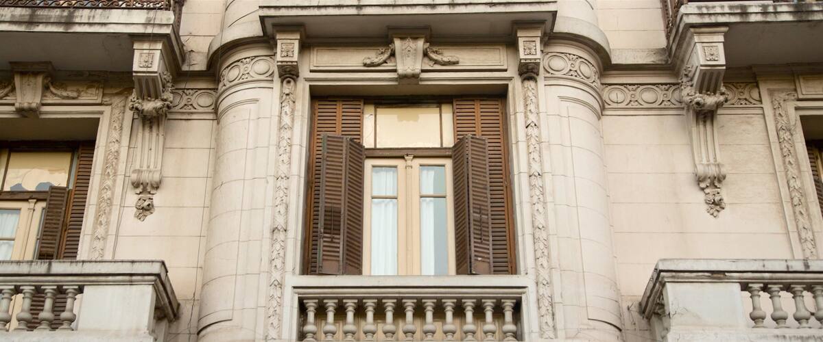 A building with ornate columns, a balcony with balustrades, and a window with wooden shutters.