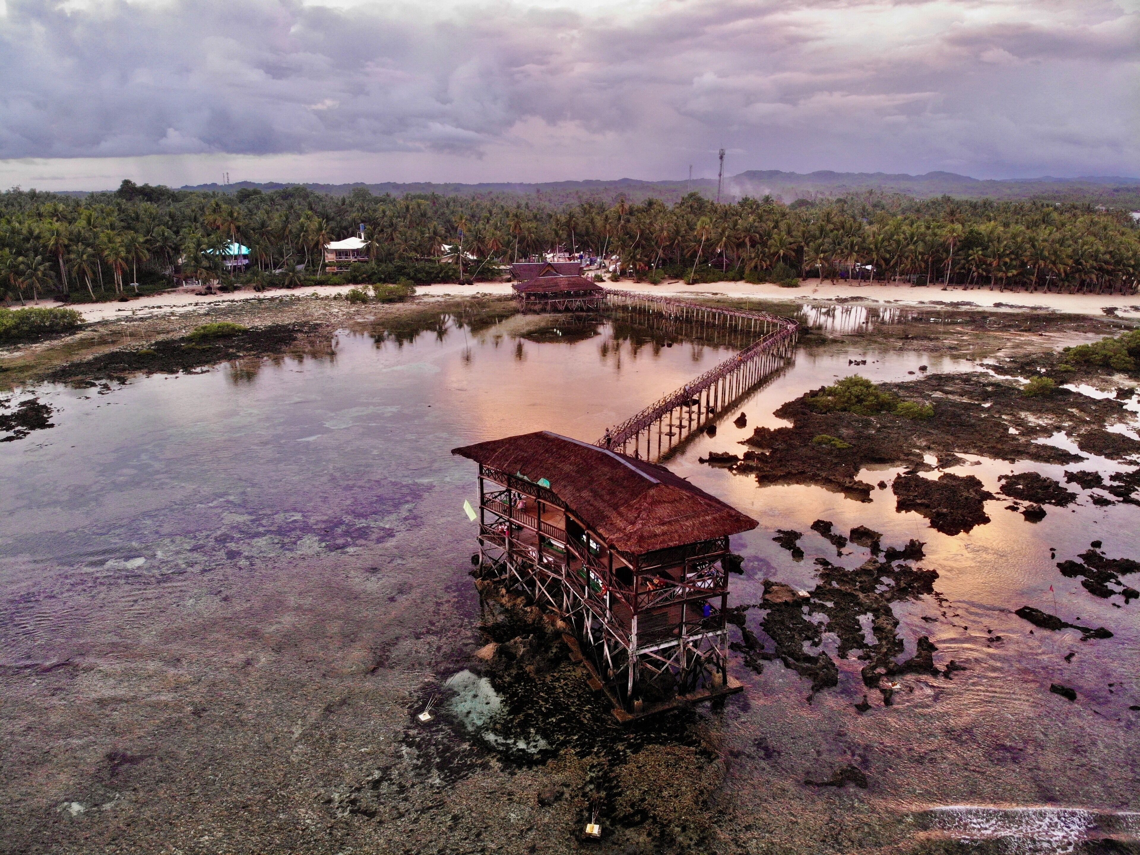 A wooden structure on stilts over water, surrounded by palm trees and a cloudy sky.